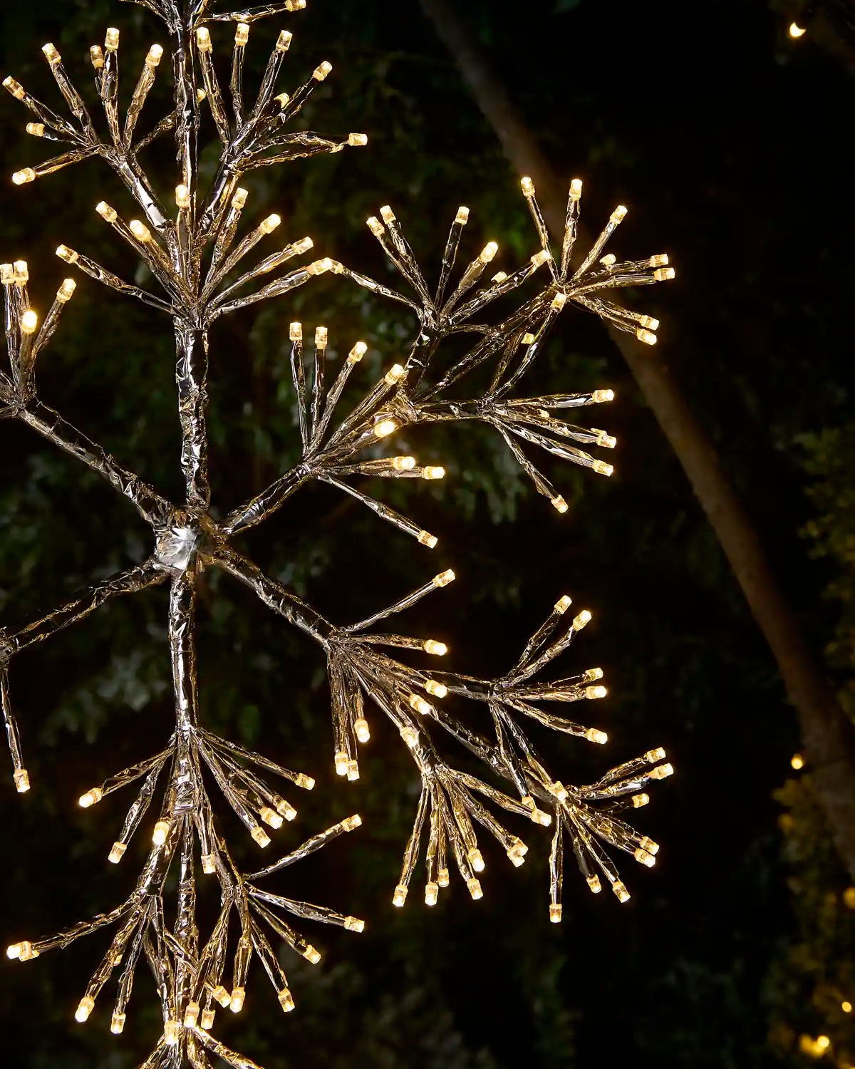 A decorative tree branch light sculpture covered in warm white LED fairy lights, photographed against a dark nighttime background with blurred trees.