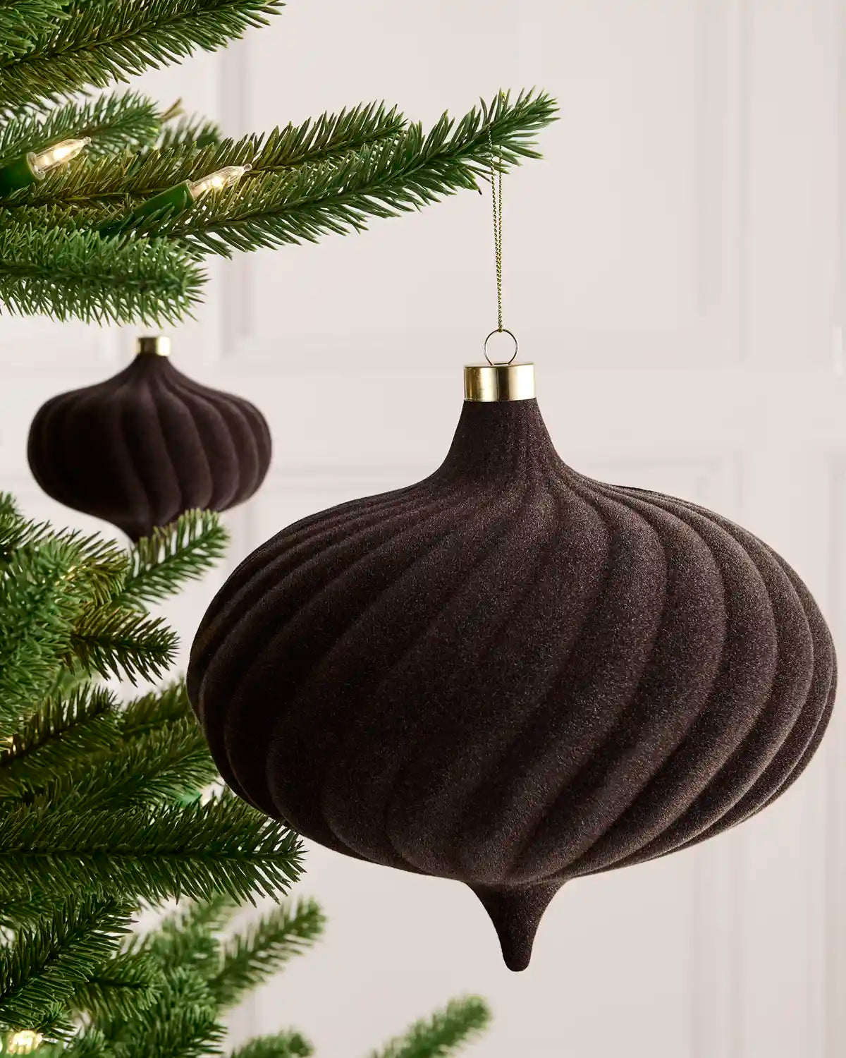 A espresso ribbed felt Christmas ornament hanging from a gold cap, suspended next to green Christmas tree branches with warm string lights.