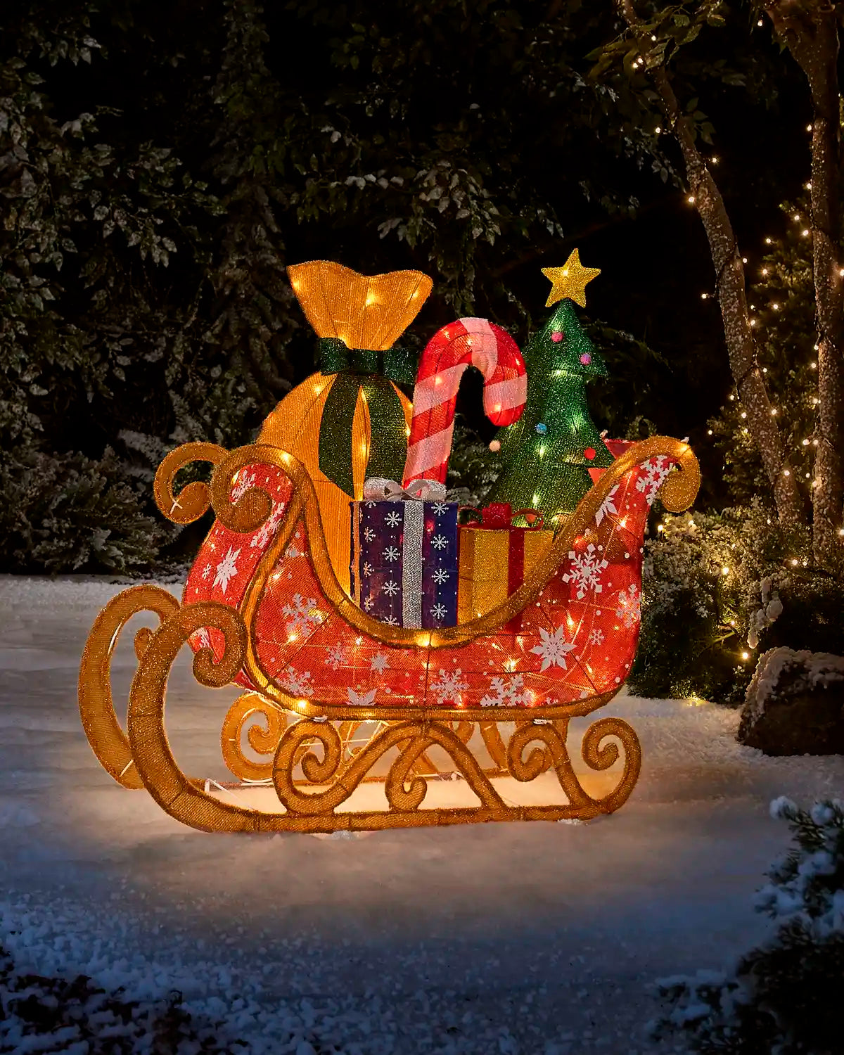 A festive Christmas-themed illuminated sleigh decoration displayed outdoors in the snow at night. The sleigh has a red patterned body with white snowflakes, a gold frame and sleigh base, a glowing green Christmas tree, candy canes, wrapped gifts, and a large gold sack with a green bow.