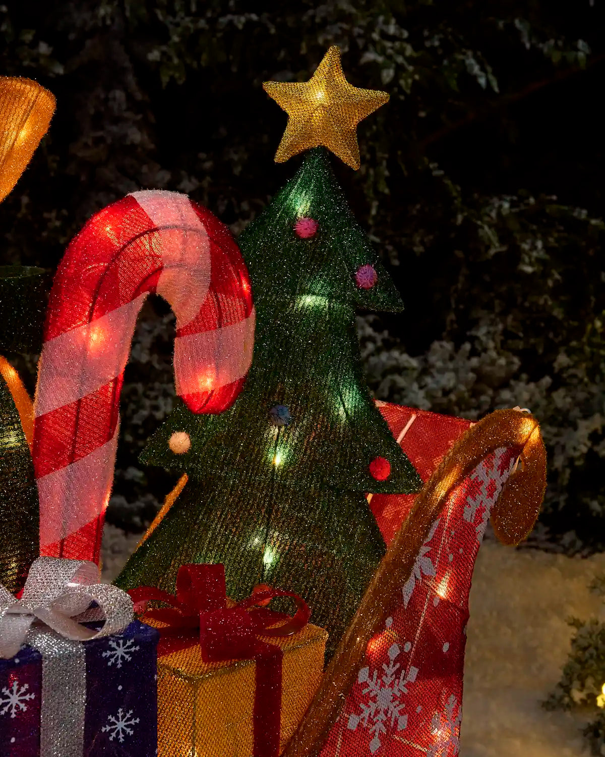 A festive Christmas decoration set featuring a green glittery tree with a gold star topper, a red and white striped candy cane, wrapped presents, and a red sleigh with gold trim and warm white string lights, set against a dark outdoor background.