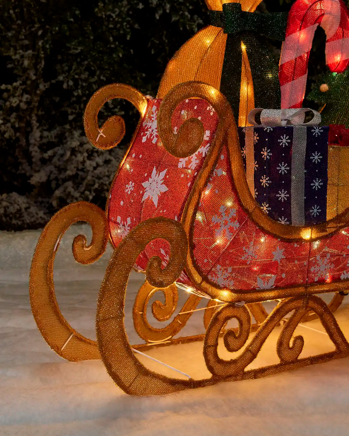 A festive Christmas sleigh decoration with gold-toned scrollwork, red and navy patterned fabric, warm white string lights, and wrapped gift details, photographed outdoors on snow at night.