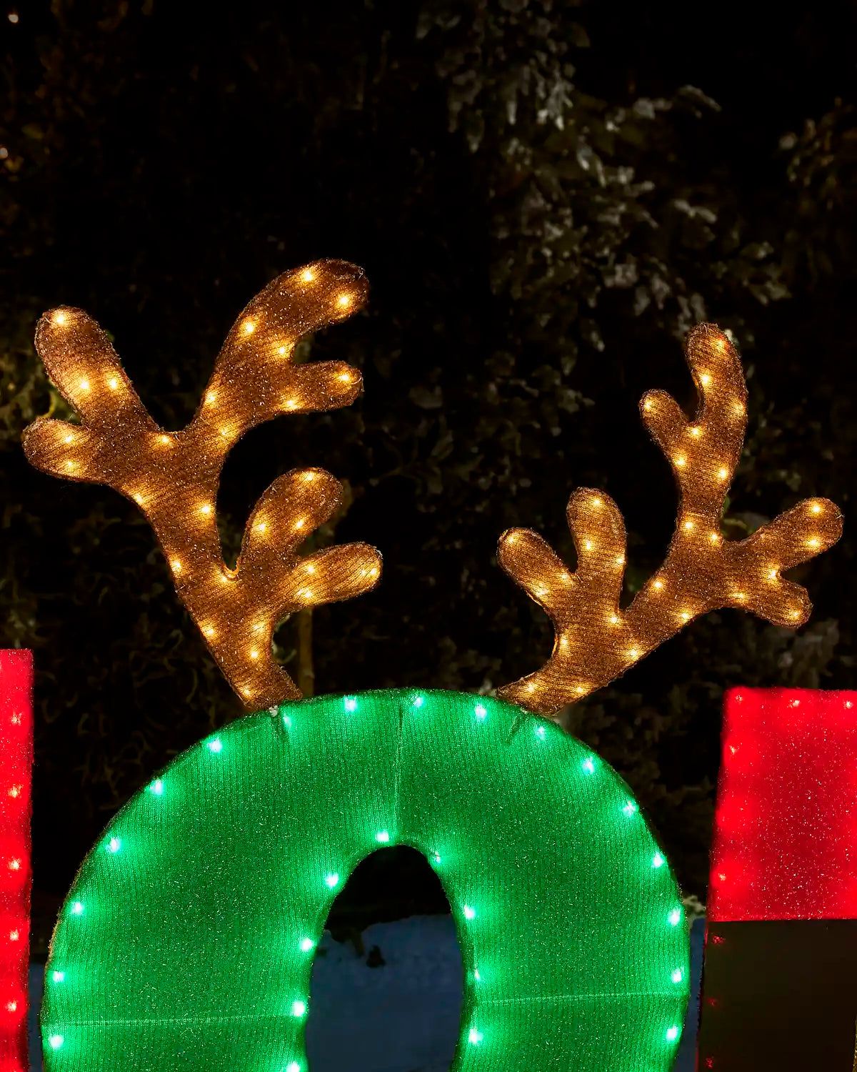 A festive holiday decoration featuring a large green oval light with a reindeer antler design on top, lit with warm yellow lights against a dark outdoor background.
