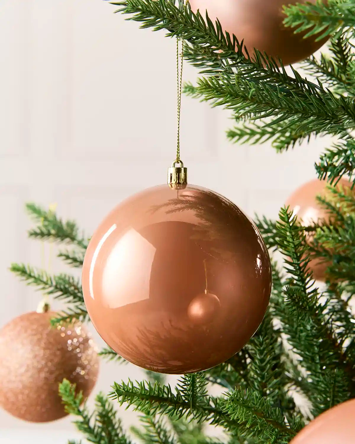A glossy metallic rose gold Christmas tree ornament hanging from a green pine branch, with other matching ornaments partially visible in the background.