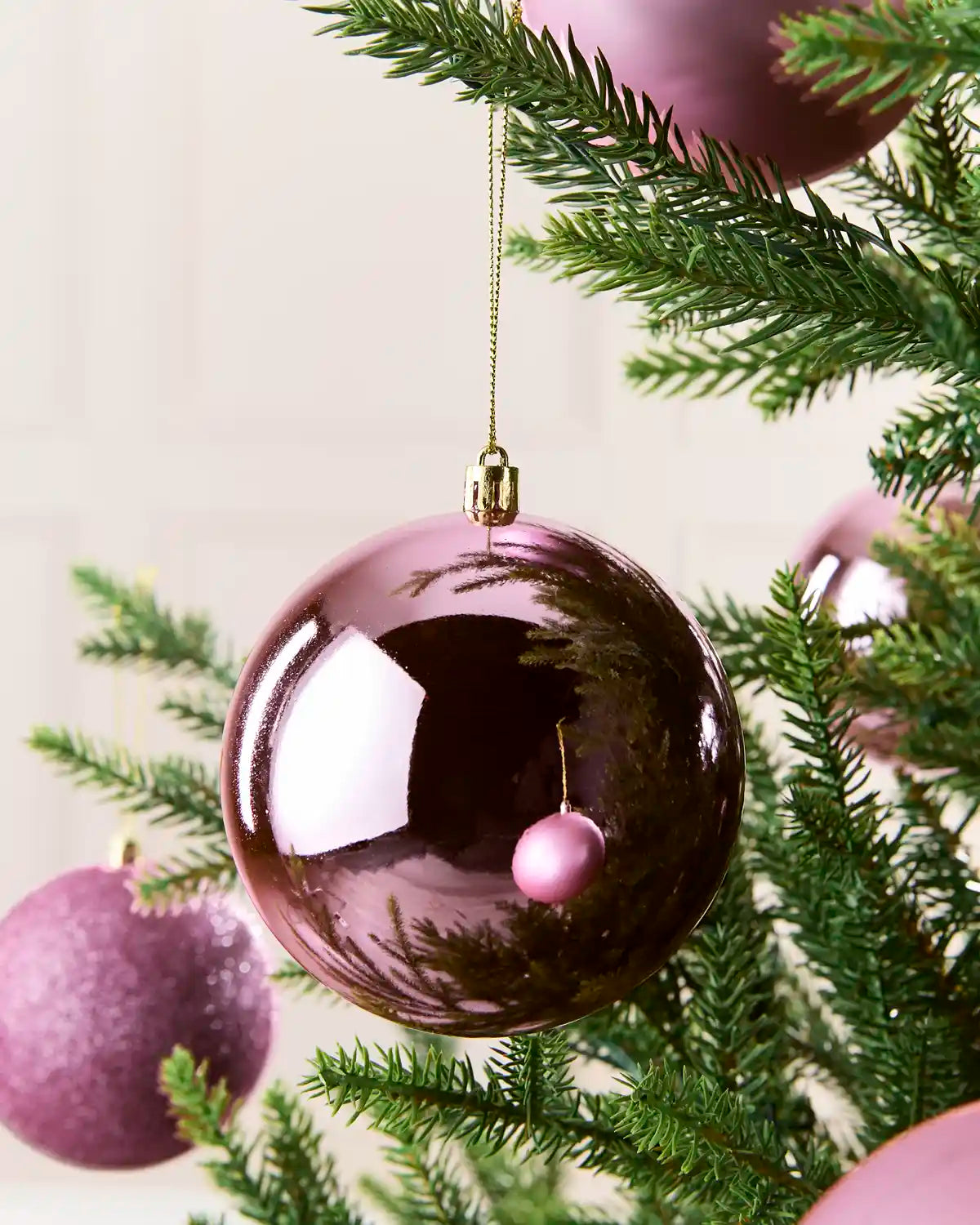A glossy, pink Christmas bauble ornament hanging from a gold string on a green Christmas tree, with other matching pink ornaments visible in the background.