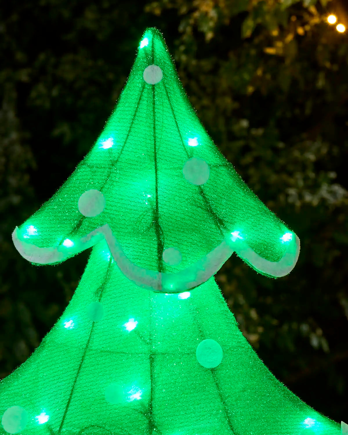A glowing green Christmas tree light made of mesh fabric with white circular ornaments and string lights, photographed outdoors against a dark, blurred background.