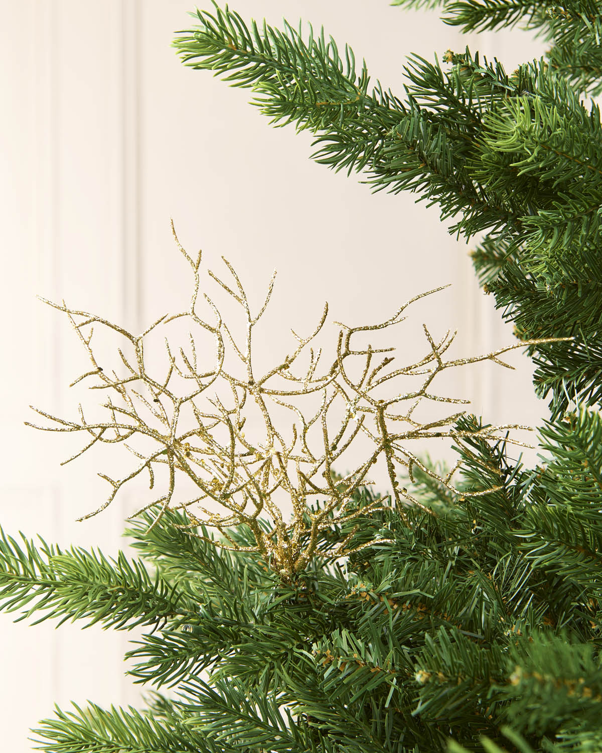 A gold glittery Coral style Christmas tree topper nestled among the green needles of an artificial pine tree, set against a soft cream-colored paneled wall background.