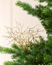 A gold glittery Coral style Christmas tree topper nestled among the green needles of an artificial pine tree, set against a soft cream-colored paneled wall background.