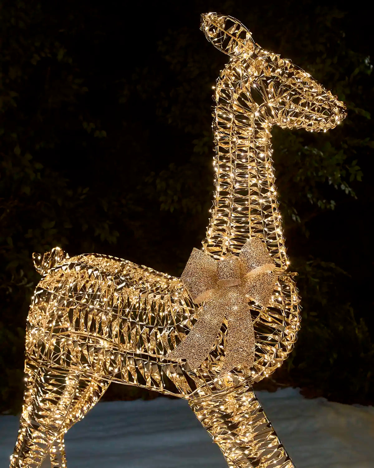 A golden wire-frame doe holiday light decoration with a glittery gold bow, shown illuminated against a dark outdoor night background.