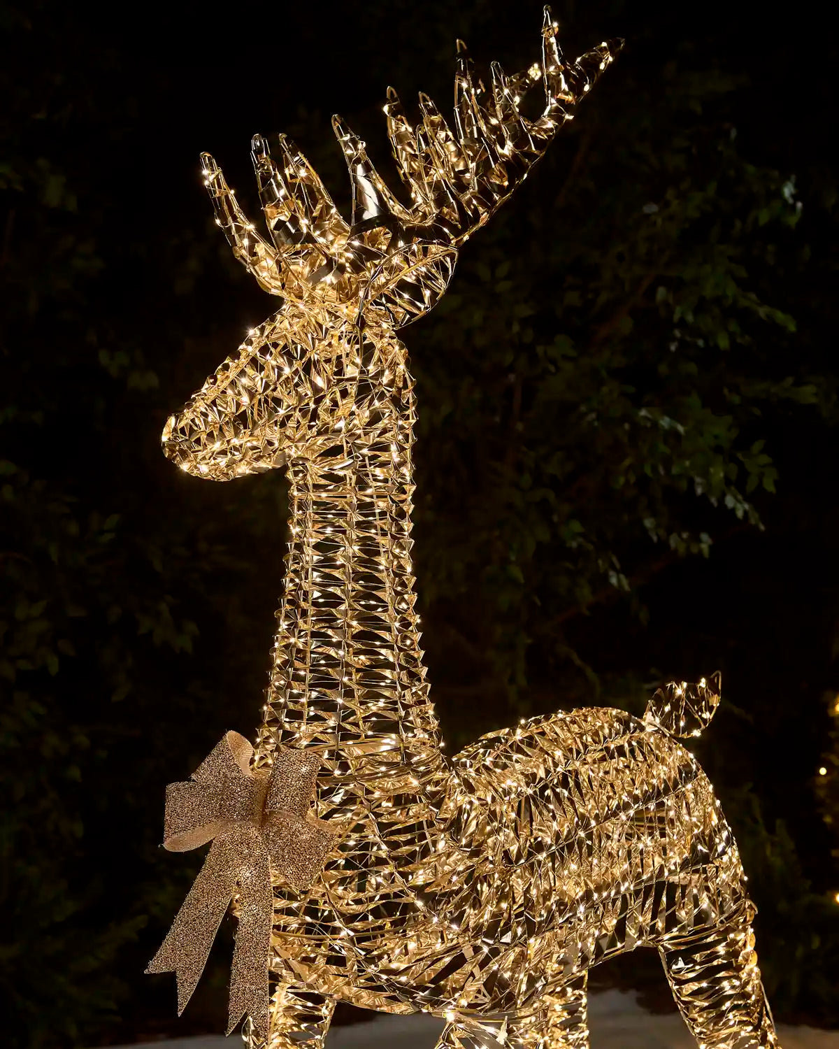 A golden wire-frame reindeer holiday light sculpture with a glittery gold bow, photographed at night against a dark, leafy outdoor background.