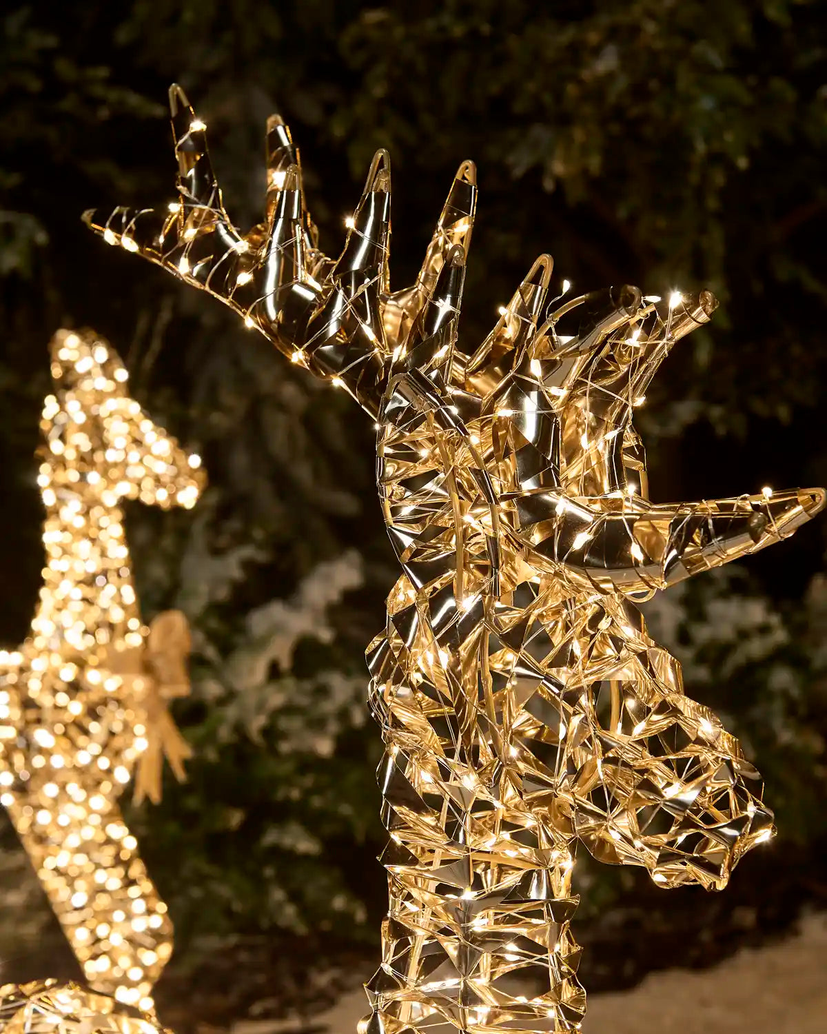 A golden wire deer head and neck light sculpture illuminated with warm white lights, photographed outdoors at night against a dark blurred background.