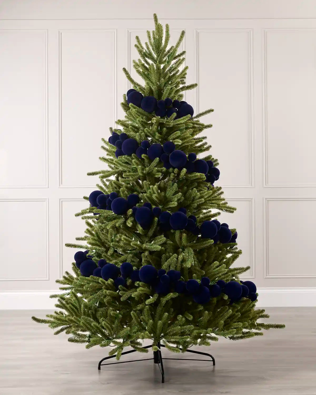 A green Christmas tree decorated with dark blue velvet baubles, standing on a black metal stand against a white paneled wall and light wood floor.