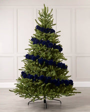 A green Christmas tree decorated with dark blue velvet baubles, standing on a black metal stand against a white paneled wall and light wood floor.