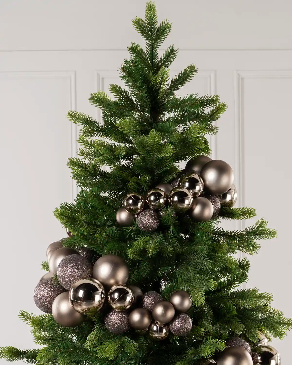 A green Christmas tree decorated with metallic Mocha coloured and glittery taupe ornaments against a light gray paneled wall.