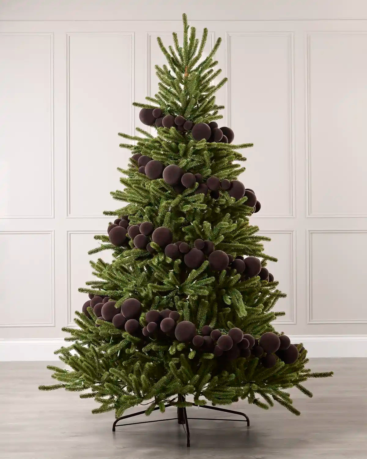 A green Christmas tree decorated with numerous Espresso coloured spherical ornaments, standing on a black metal stand against a white paneled wall and light gray floor.