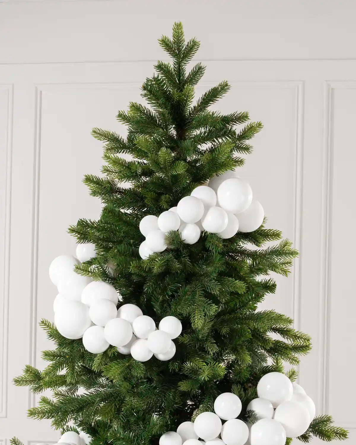 A green artificial Christmas tree decorated with clusters of white balloons, set against a white paneled wall background.