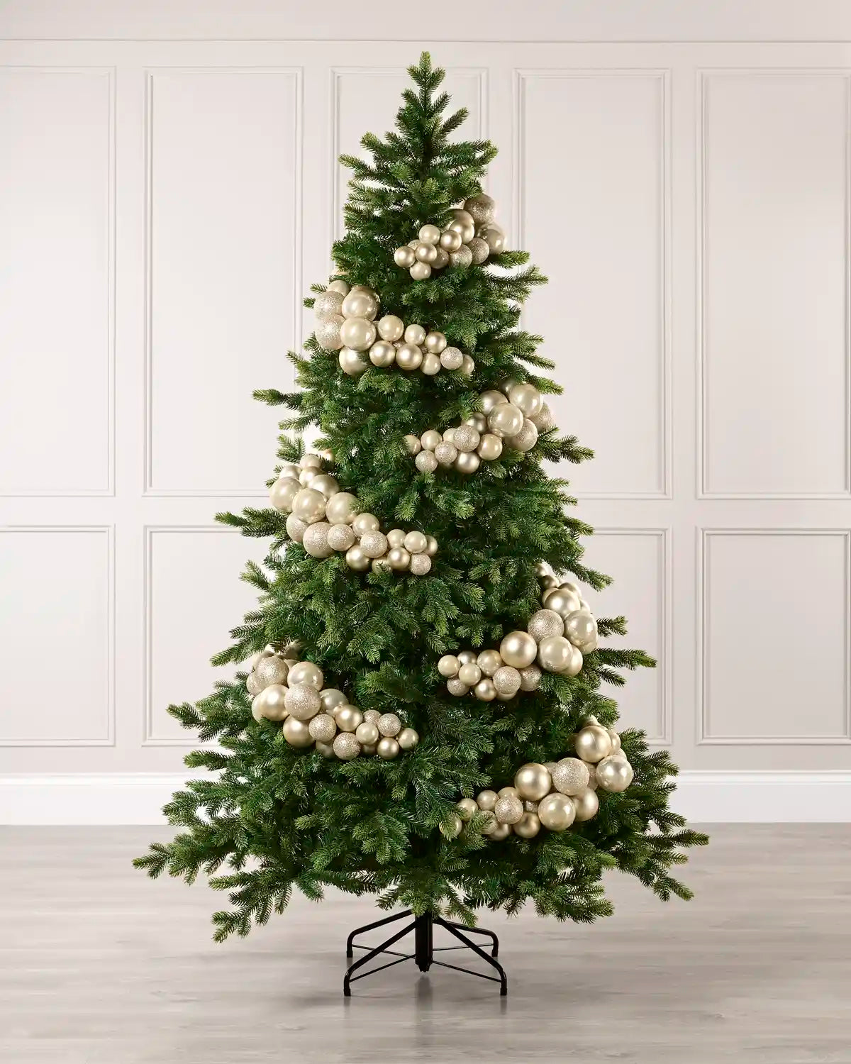 A green artificial Christmas tree decorated with large Latte spherical ornaments, standing on a black metal stand against a white paneled wall and light wood floor.