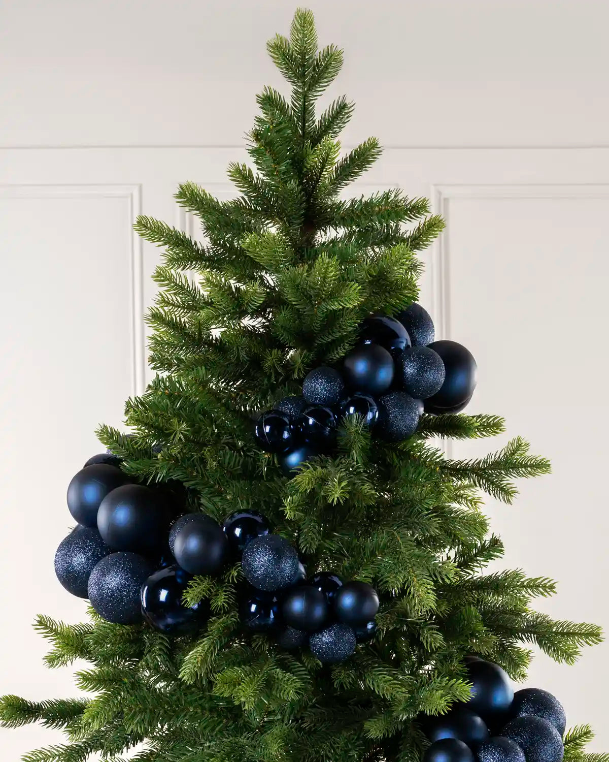 A green artificial Christmas tree decorated with matte and glittery dark blue spherical bauble garland arch ornaments, set against a light beige paneled wall.