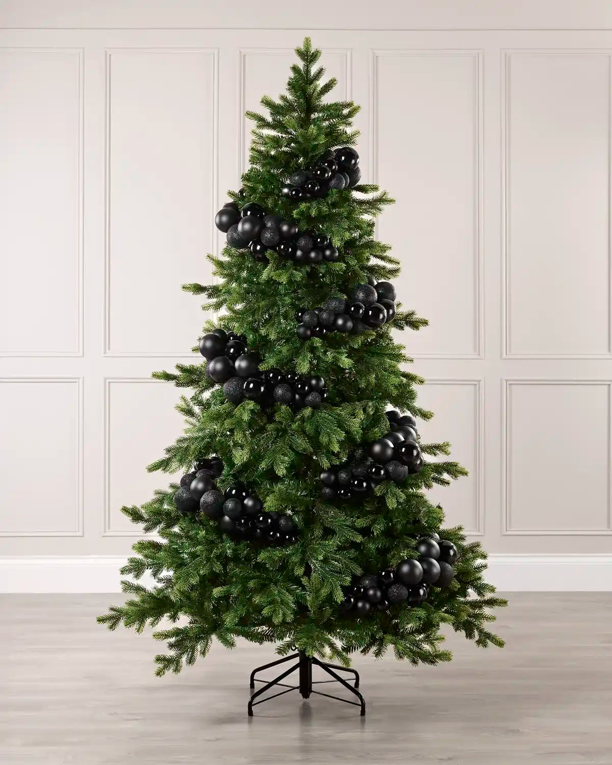 A green artificial Christmas tree decorated with numerous glossy black spherical ornaments, standing on a black metal stand against a light beige paneled wall and light wood floor.