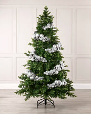 A green artificial Christmas tree decorated with numerous shiny silver ornaments bauble arch garland, standing on a black metal stand against a light beige paneled wall and light wood floor.