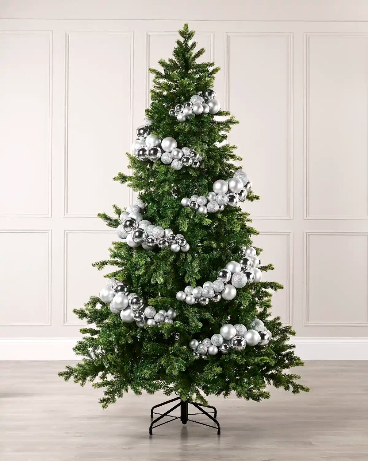 A green artificial Christmas tree decorated with numerous shiny silver ornaments bauble arch garland, standing on a black metal stand against a light beige paneled wall and light wood floor.