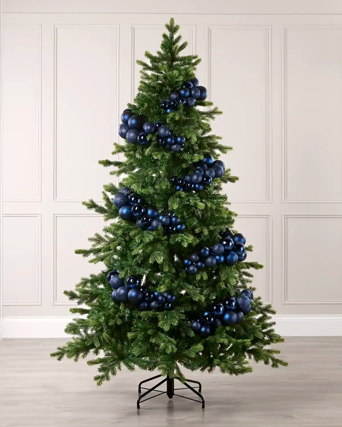 A green artificial Christmas tree decorated with shiny blue and navy spherical ornaments, standing on a black metal stand against a light beige paneled wall and wood floor.