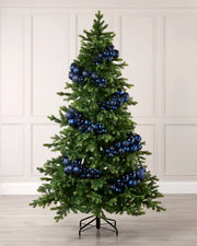 A green artificial Christmas tree decorated with shiny blue and navy spherical ornaments, standing on a black metal stand against a light beige paneled wall and wood floor.