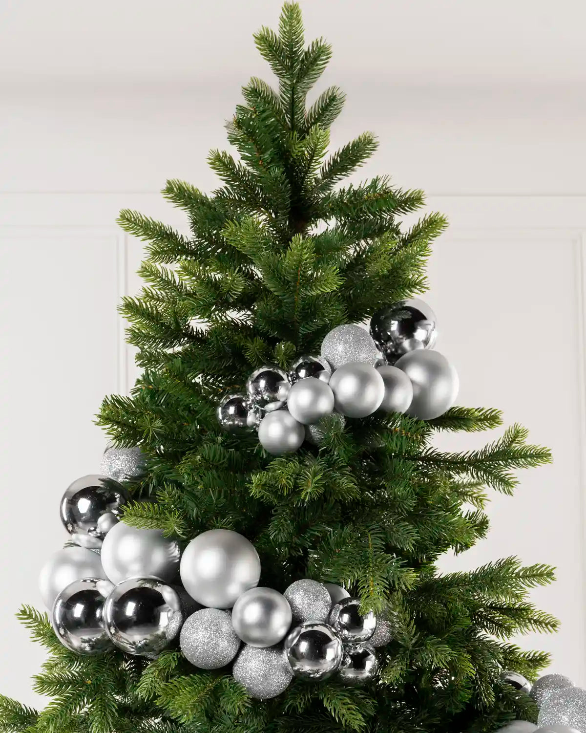 A green artificial Christmas tree decorated with shiny silver and matte baubles garland arch ornaments, shown against a plain light wall.