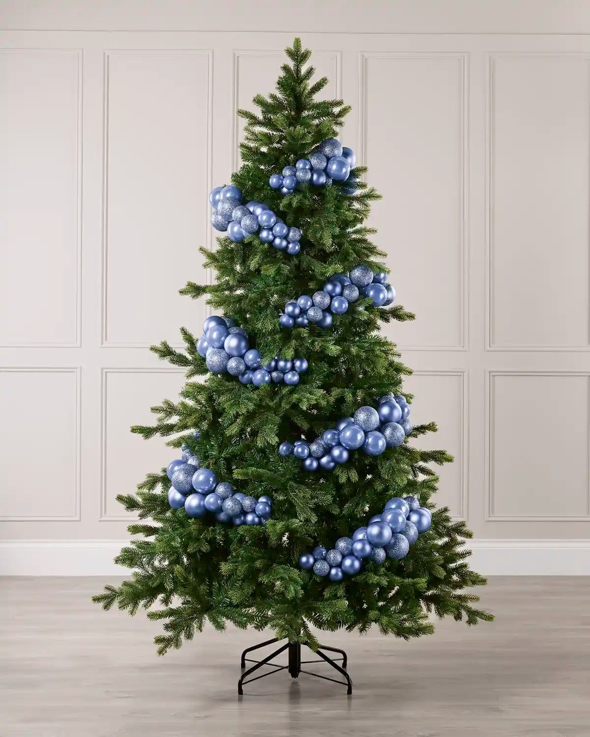 A green artificial Christmas tree with Ice blue metallic ball garlands, displayed on a black stand against a light beige paneled wall and wood floor.