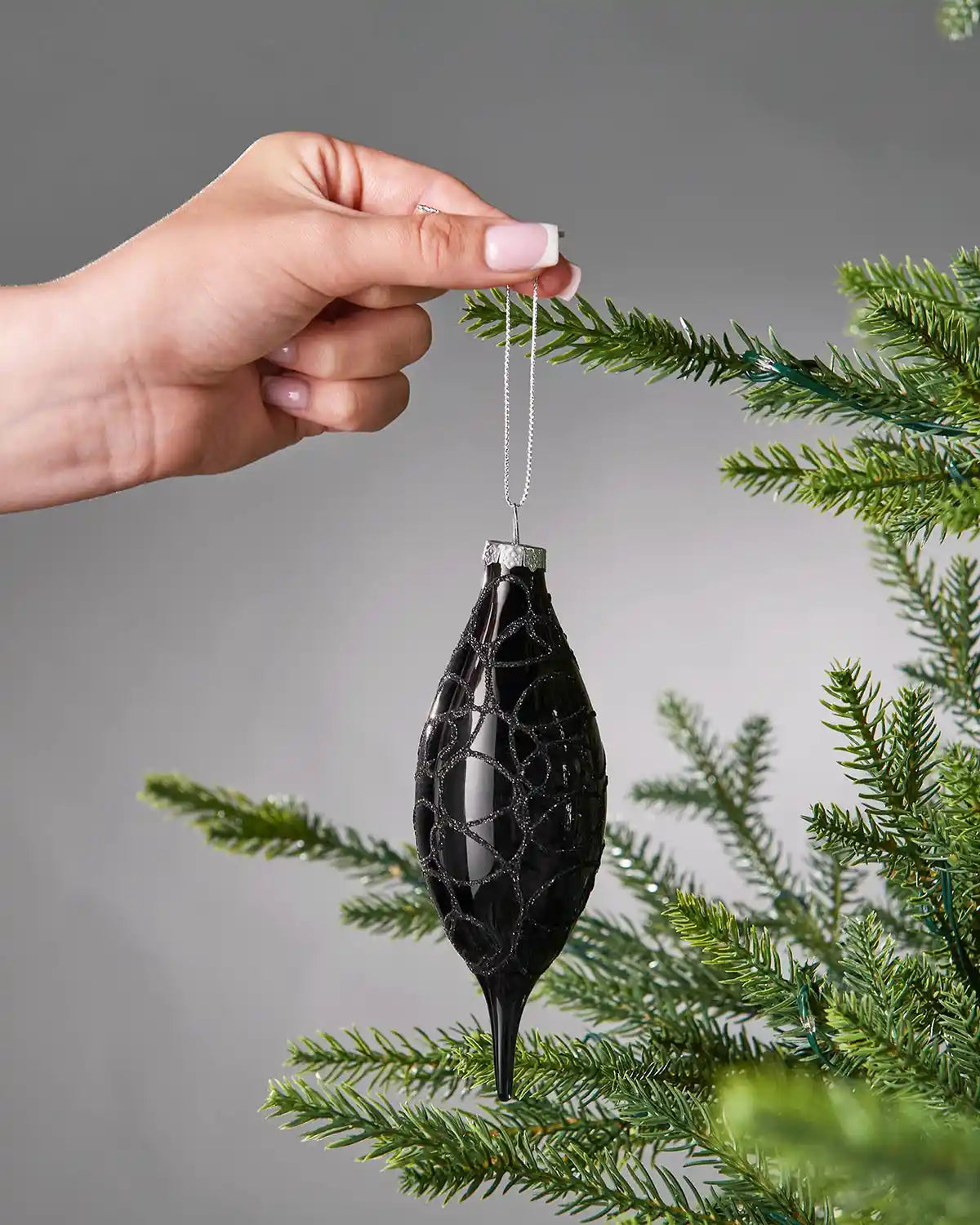 A hand holding a Black decorative ornament in front of a tree branch against a grey background