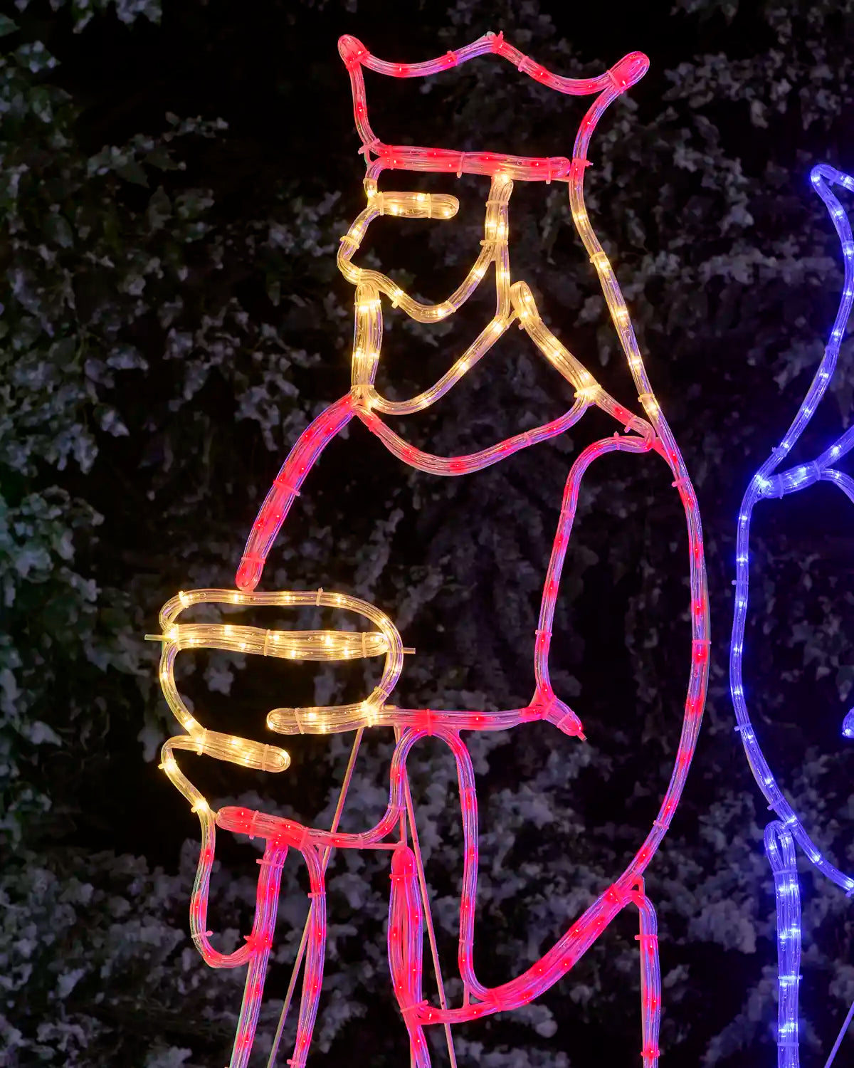A large Christmas light decoration shaped like a crowned figure, outlined with red LED lights and filled with warm yellow lights, photographed outdoors at night against dark foliage. Part of a blue light decoration is visible on the right edge.