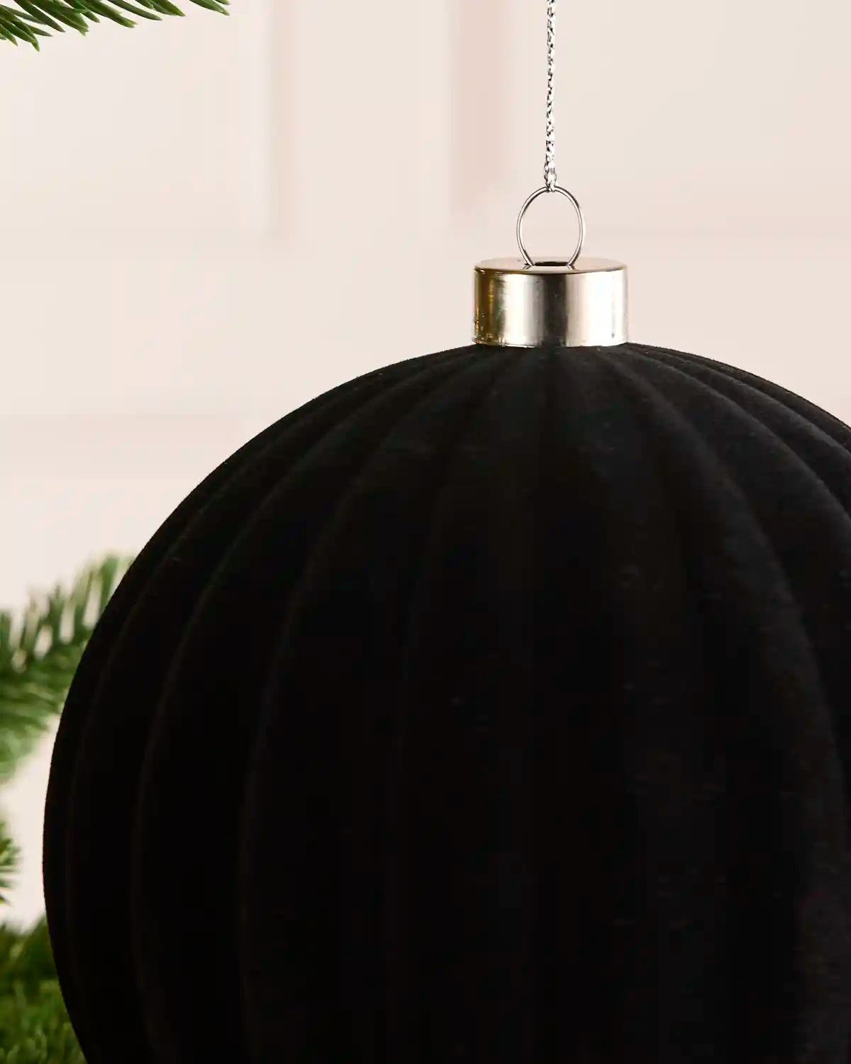 A large black ribbed hanging ornament with a metallic gold cap and silver hanging chain, shown close up against a soft light background with green pine branches at the edges.