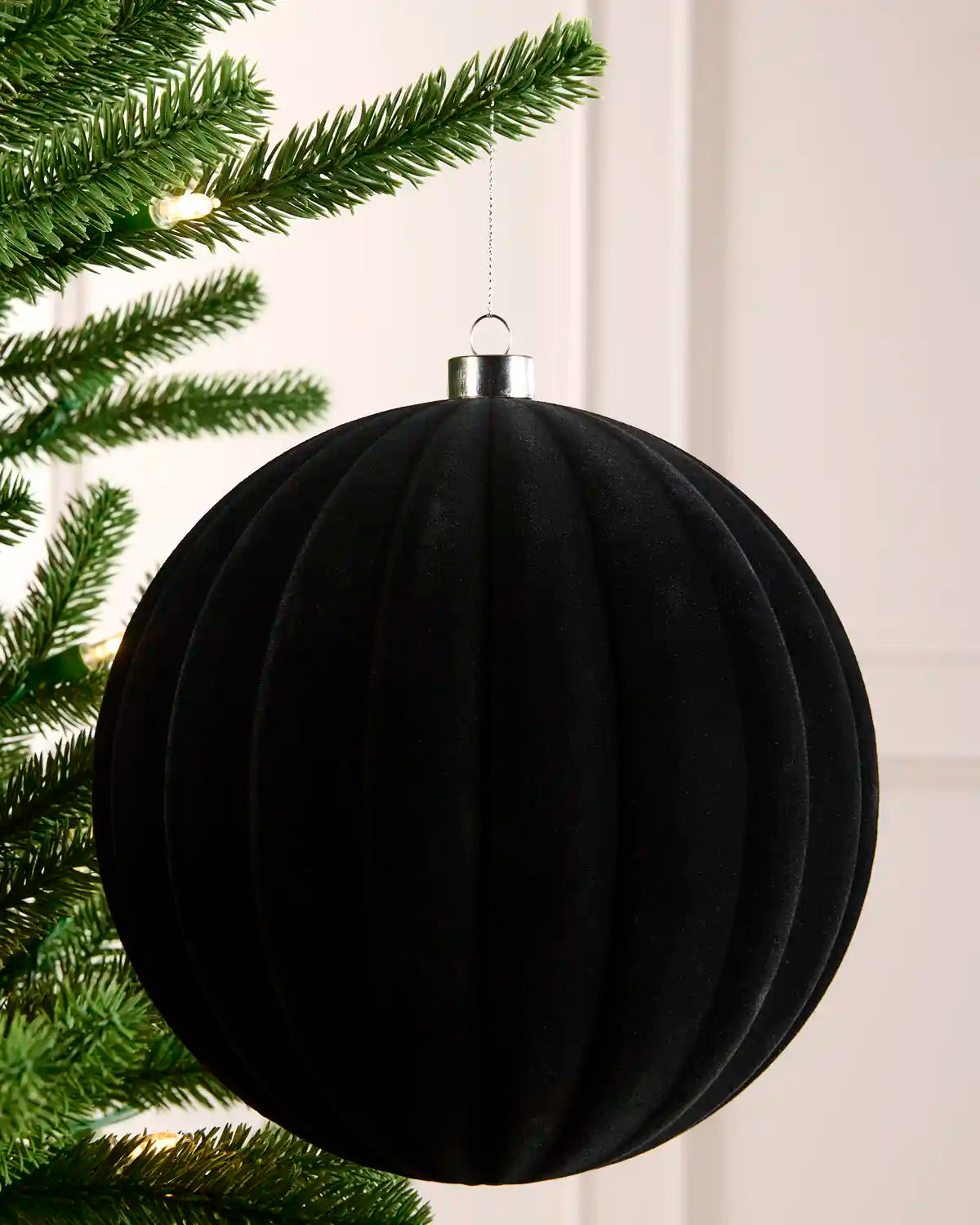 A large black ribbed velvet Christmas ornament hanging from a silver cap next to green Christmas tree branches, against a light background