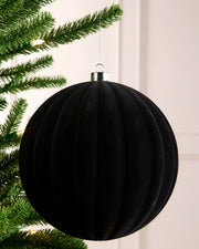 A large black ribbed velvet Christmas ornament hanging from a silver cap next to green Christmas tree branches, against a light background