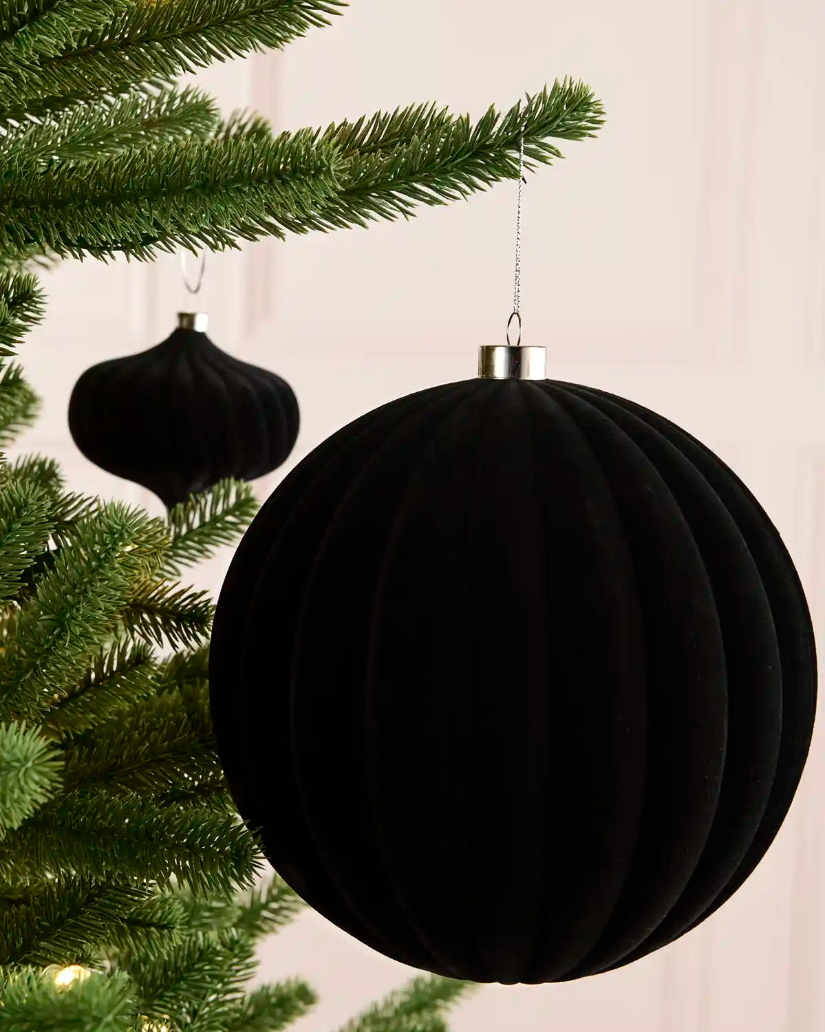 A large black ribbed velvet Christmas ornament hanging from a silver cap next to green Christmas tree branches, with a second matching ornament blurred in the background.