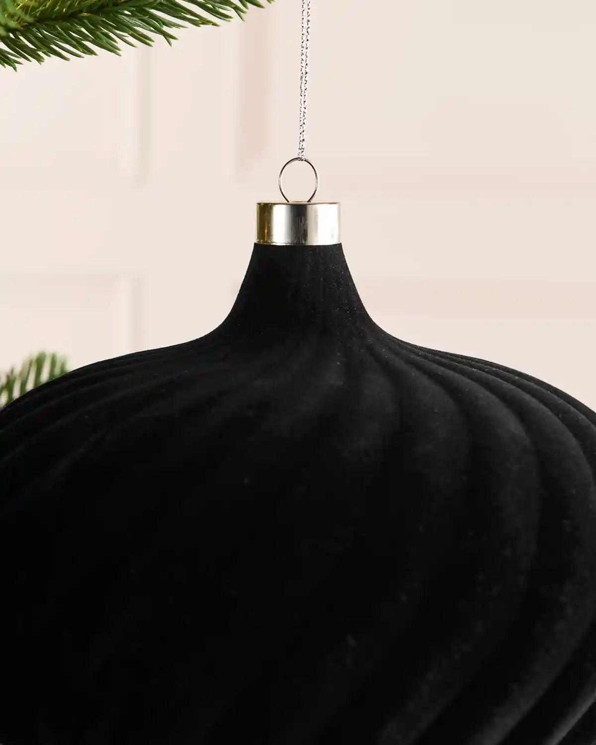 A large black ribbed velvet Christmas ornament hanging from a silver cap with a chain, shown against a soft cream background with pine branches at the top.