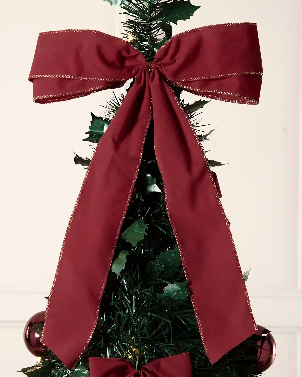A large burgundy Christmas tree topper bow with gold trim, displayed on a green artificial Christmas tree.