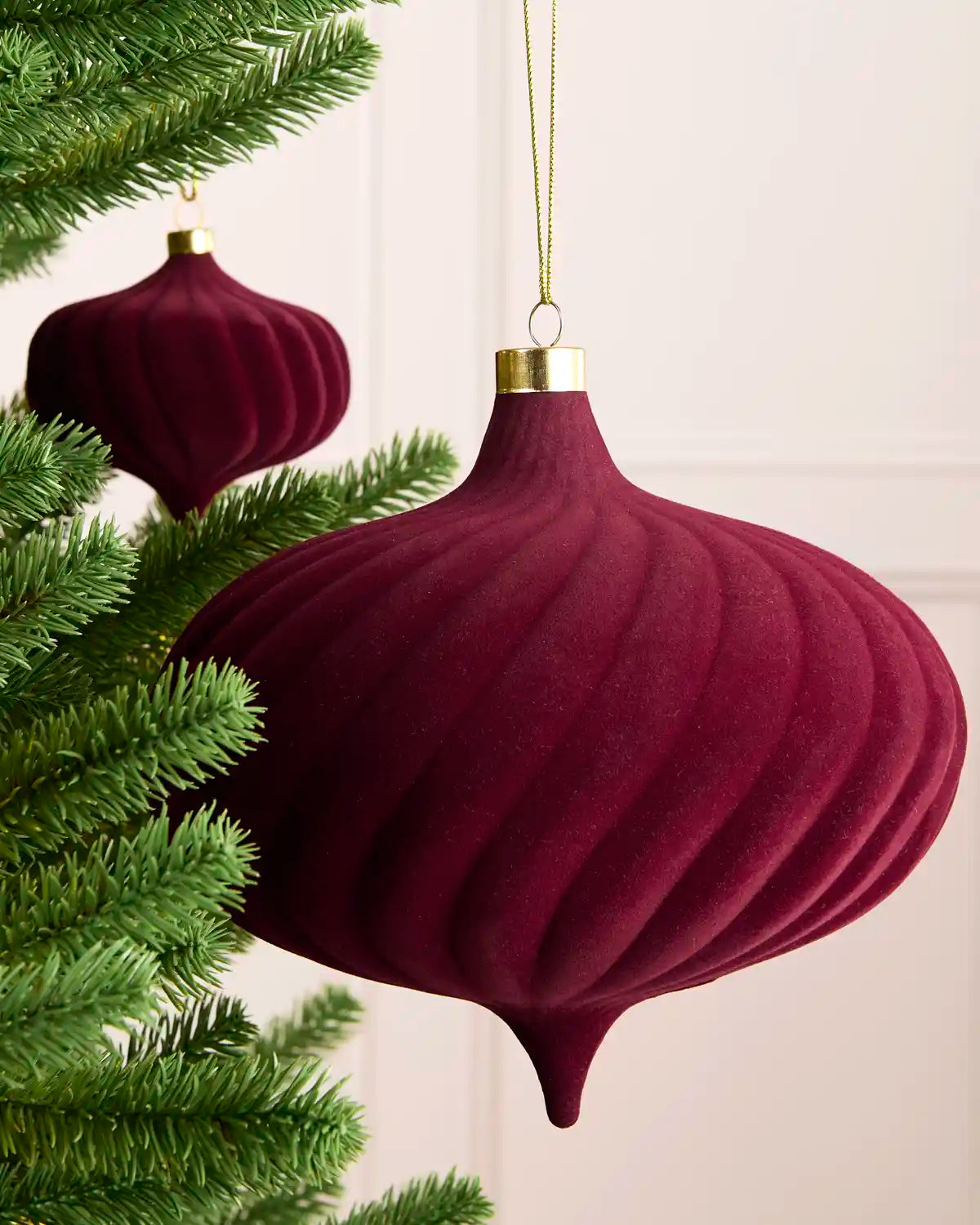 A large burgundy velvet Christmas ornament shaped like an onion, hanging from a gold string next to green pine tree branches against a light cream wall.