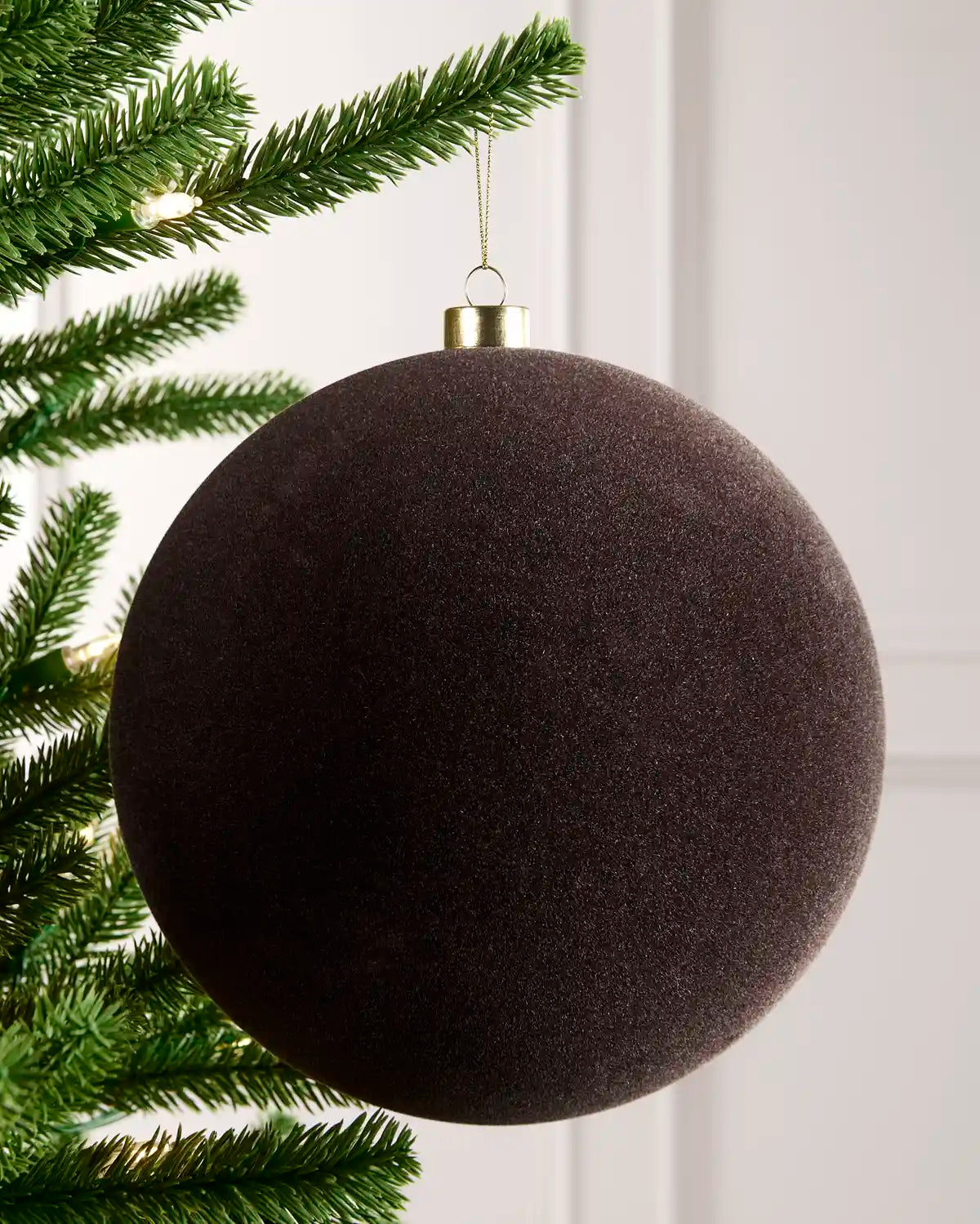 A large, espresso coloured textured spherical Christmas ornament hanging from a gold cap on a green artificial Christmas tree branch against a white panelled wall.