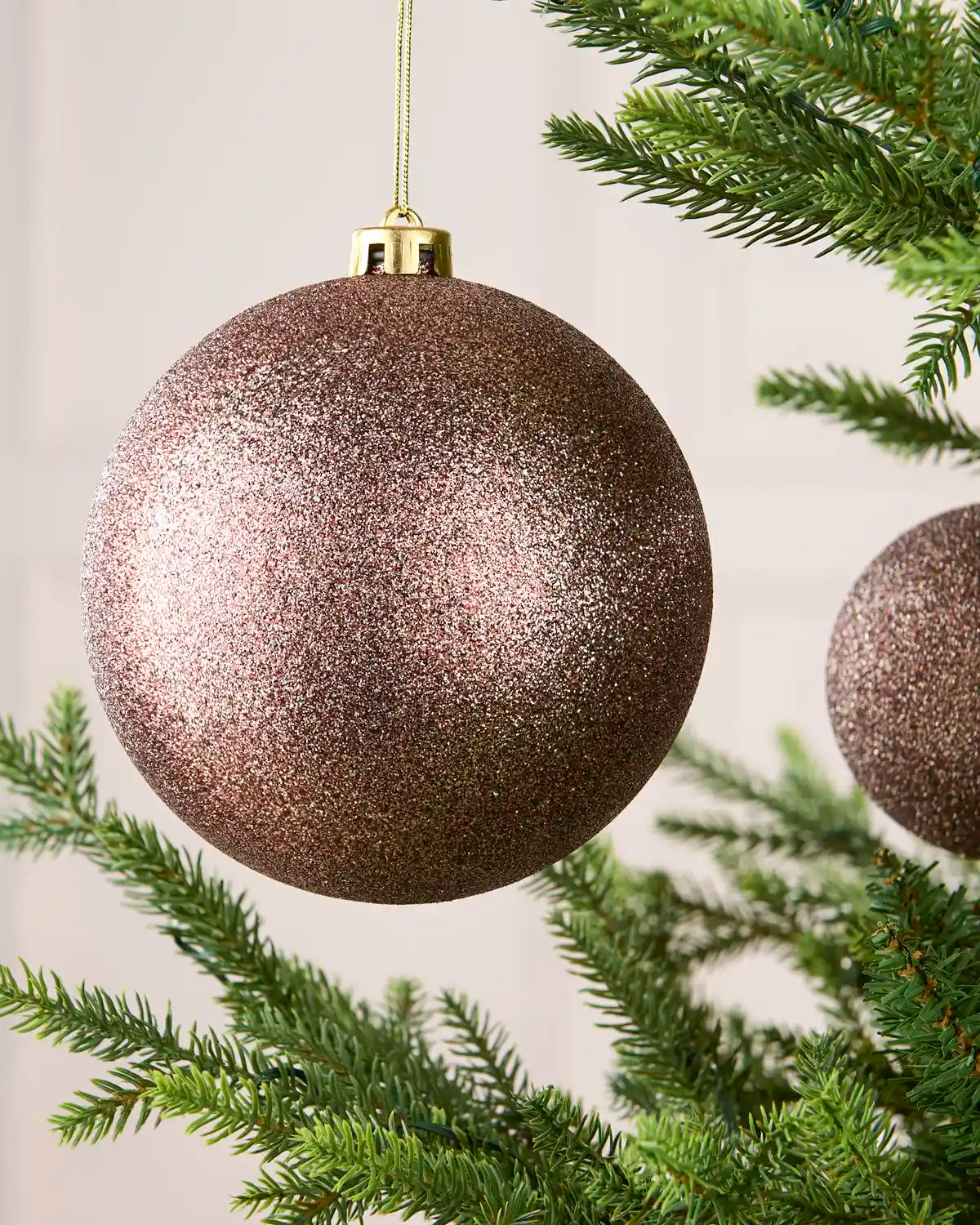 A large glittery Mocha Christmas ornament hanging from a gold cap on a tree branch, with green pine needles and part of another ornament visible in the background.