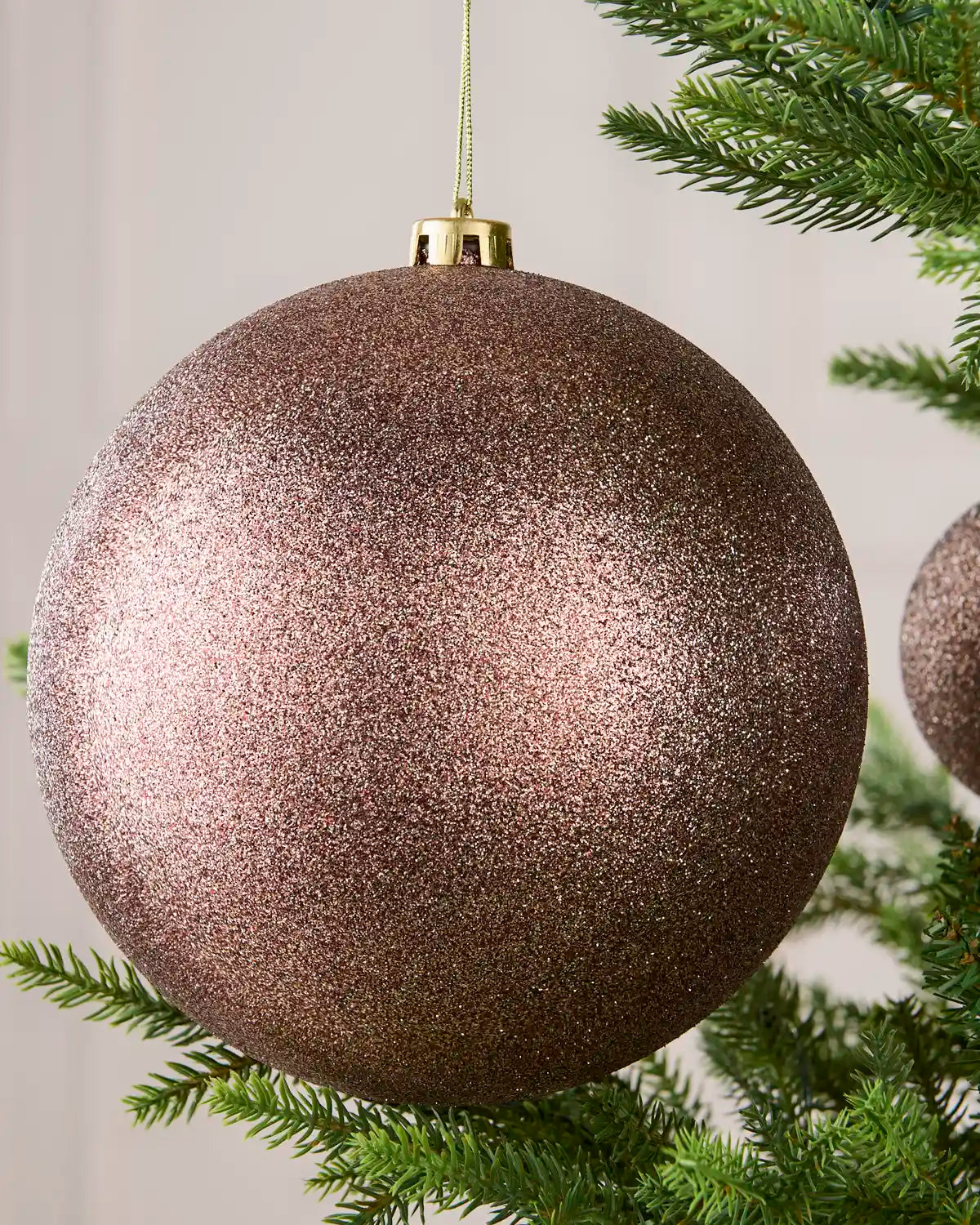 A large glittery Mocha Christmas ornament hanging from a green pine tree branch against a soft neutral background.
