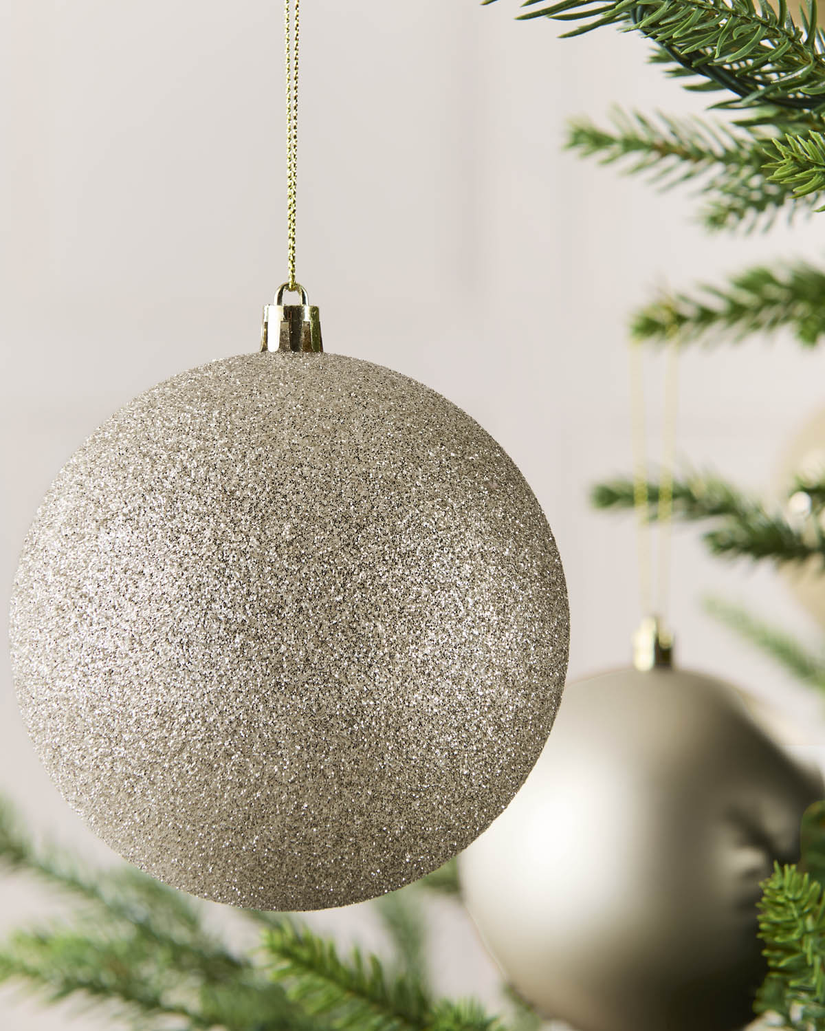 A large glittery latte Christmas tree ornament hanging from a gold string, with pine branches and a softly blurred second ornament in the background.