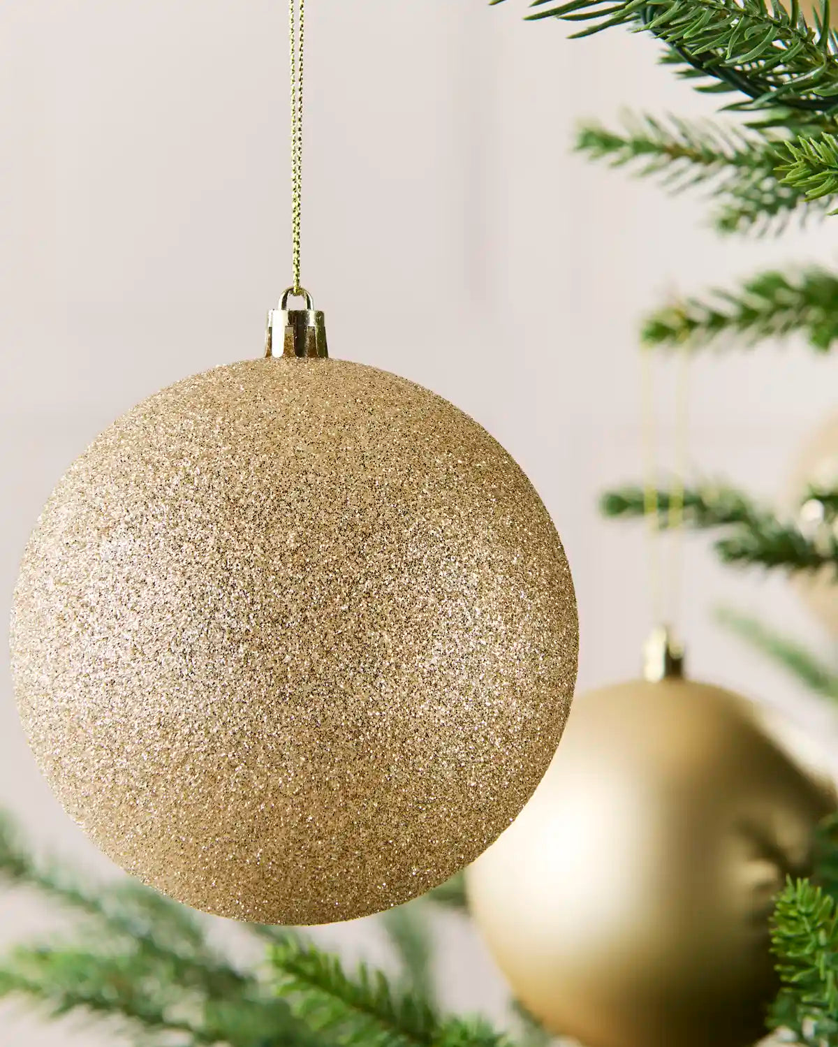 A large glittery latte Christmas tree ornament hanging from a gold string, with pine branches and a softly blurred second ornament in the background.
