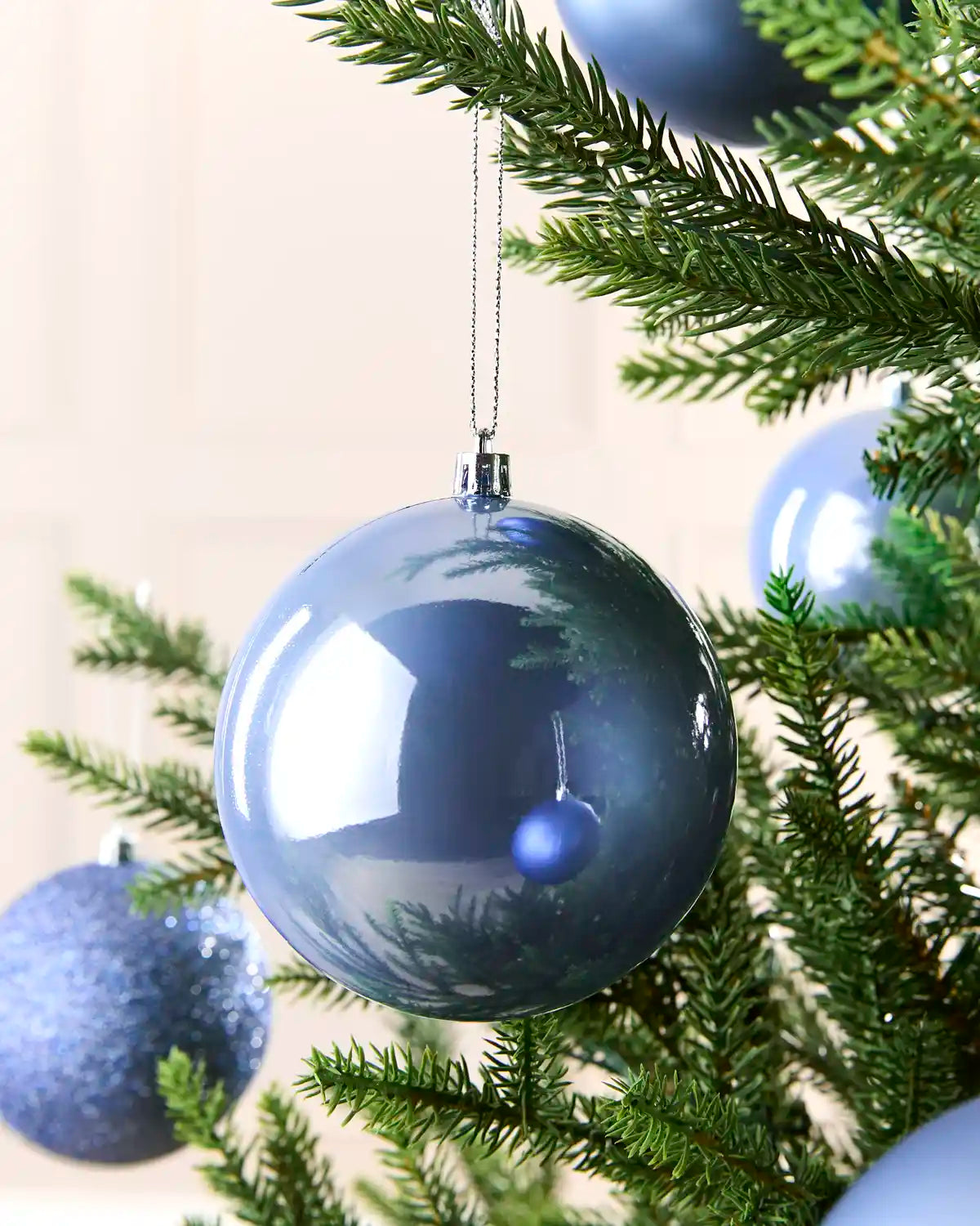 A large, glossy Ice blue Christmas bauble ornament hanging from a green pine tree branch, with other blue ornaments and a soft beige background visible.