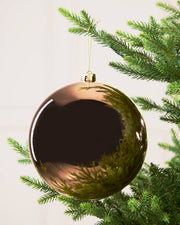 A large, glossy Mocha Christmas bauble ornament hanging from a green pine tree branch against a soft off-white background.