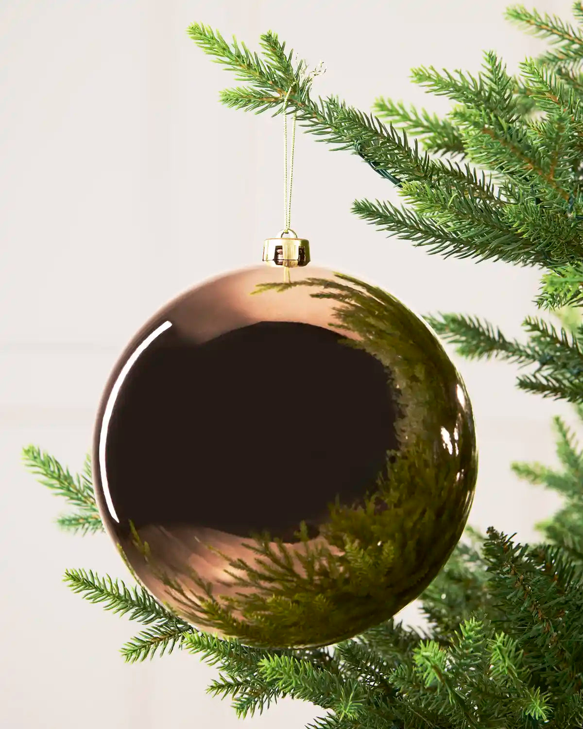A large, glossy Mocha Christmas bauble ornament hanging from a green pine tree branch against a soft off-white background.