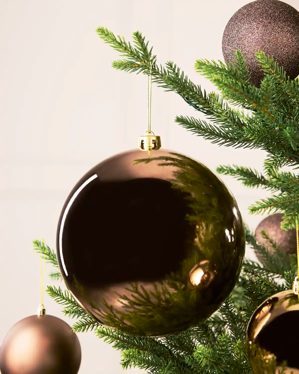 A large, glossy Mocha Christmas bauble ornament hanging from a green pine tree branch with smaller Mocha baubles behind against a soft off-white background.