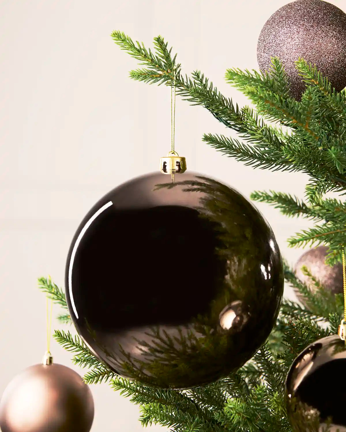 A large glossy Mocha Christmas tree ornament hanging from a green evergreen branch, with other metallic tree ornaments and a soft neutral background.