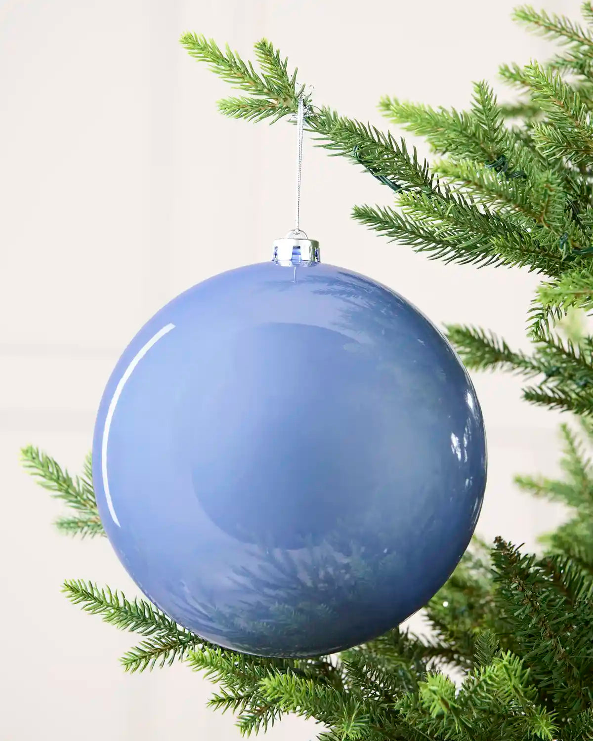 A large glossy blue Christmas ornament hanging from a green evergreen tree branch against a soft off-white background.