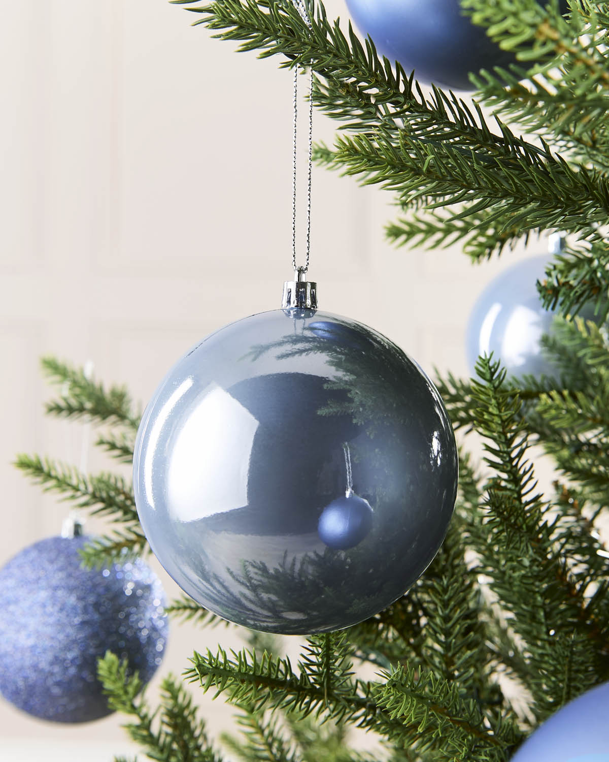 A large, glossy dark ice blue glass Christmas ornament hanging from a green pine tree branch, with other blue ornaments and the tree branches in the background.