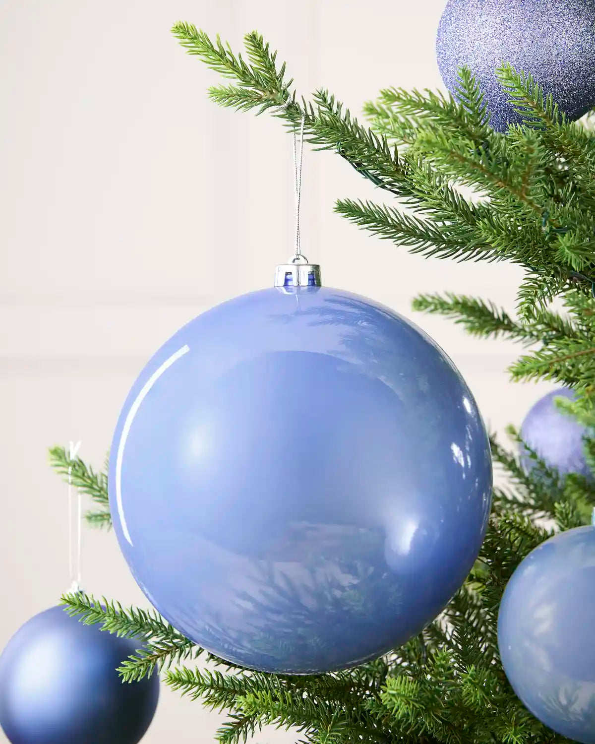 A large glossy light blue Christmas ornament hanging from a green evergreen tree branch, with other matching blue ornaments partially visible around it.